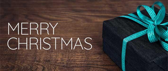 Merry Christmas with a present wrapped in black paper with a green bow sitting on a brown table