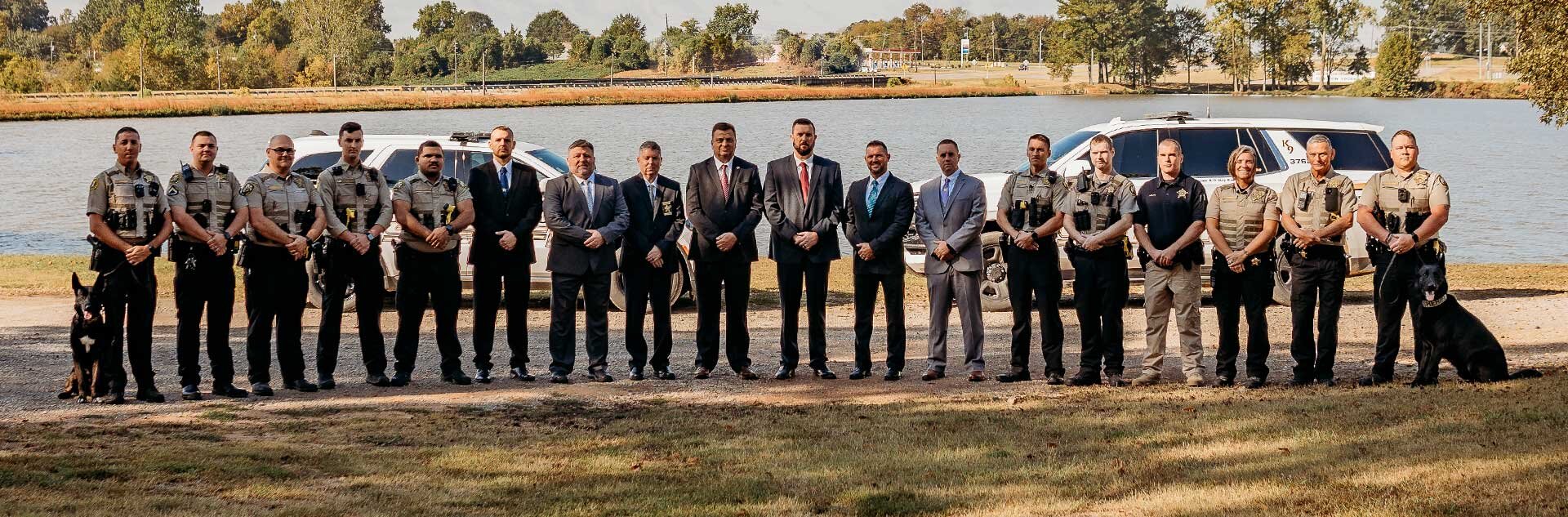 Sheriff's Office Staff stand in 1 row in front of vehicles and a lake
